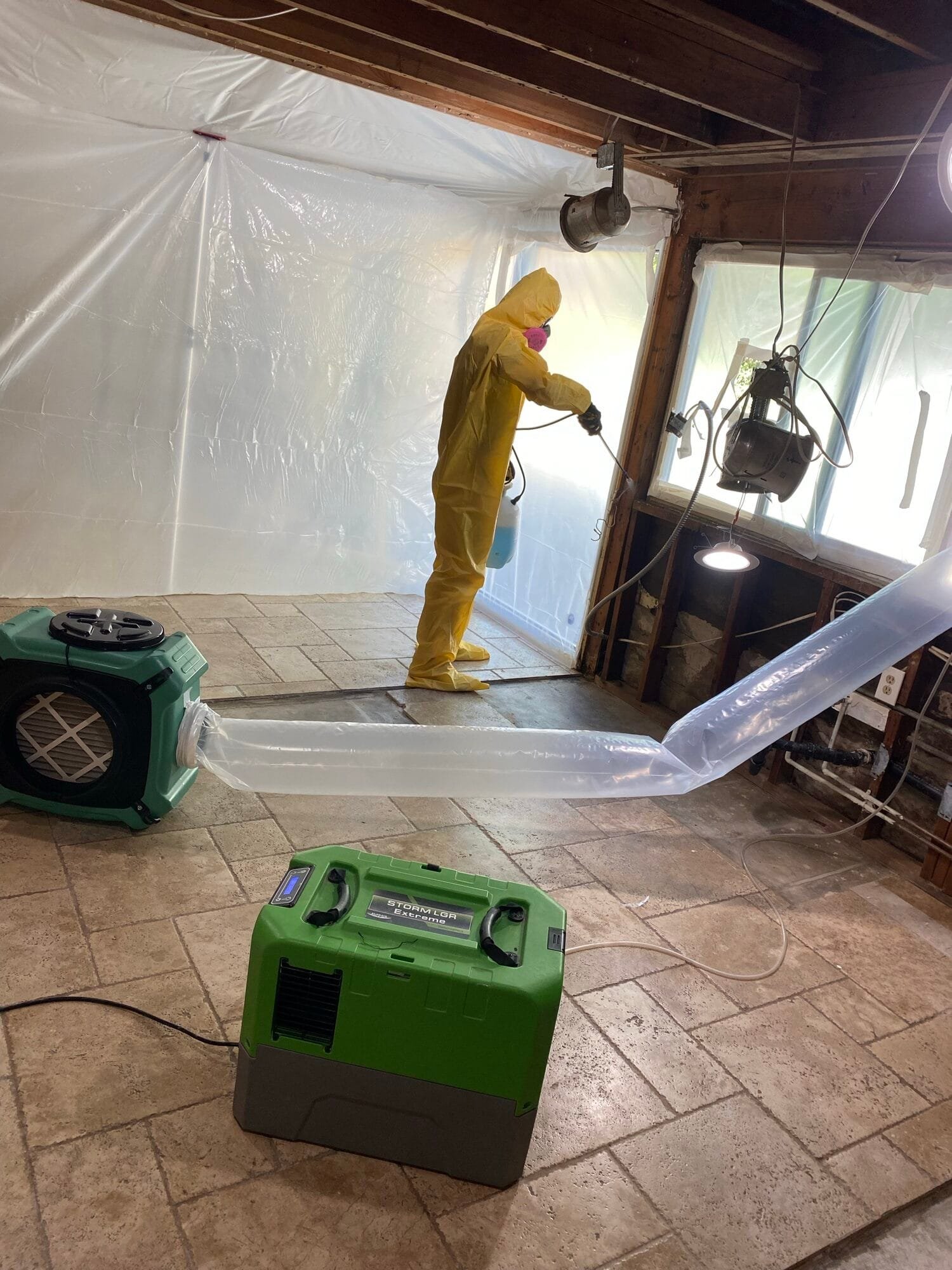 Technician in a protective suit disinfecting framing inside containment, with a HEPA air scrubber ducted outside