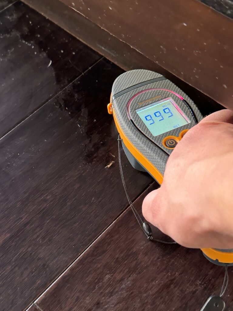 Moisture meter showing a wet reading on a wood floor next to a kitchen cabinet after a refrigerator line leak in Sherman Oaks