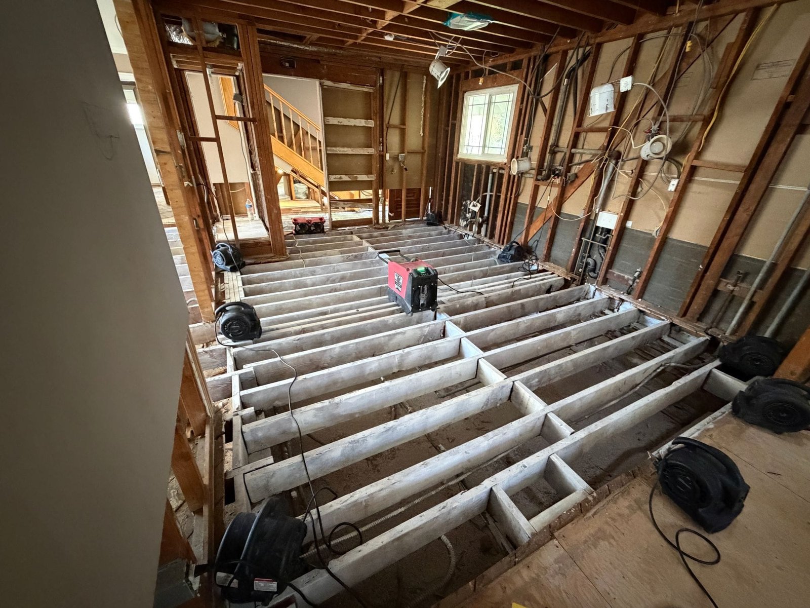 Water-damaged subfloor and drywall removed down to the joists and framing, with drying equipment in place